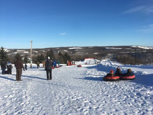 People enjoy snow tubing on a sunny winter day, with snowy hills and trees in the background.