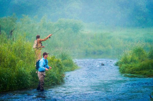 Two fishermen casting lines into a misty river surrounded by lush greenery.