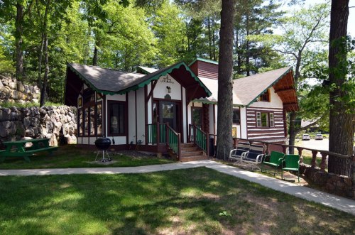 A charming cabin with a unique design, surrounded by trees and a grassy area, featuring a stone wall and outdoor seating.