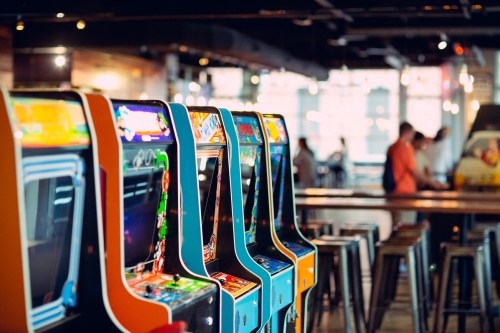 A row of colorful arcade machines in a lively gaming venue with people in the background.