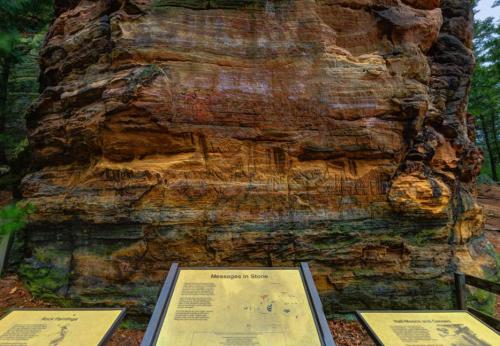 A large rock face with colorful layers and two informational signs in the foreground about geological features.