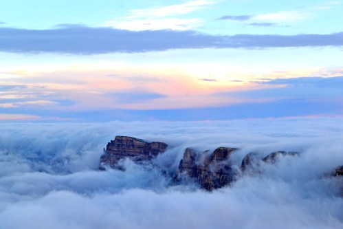 A dramatic landscape of mountains emerging from thick clouds under a colorful sky at sunset.