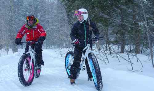 Two people riding fat bikes through a snowy forest, smiling and enjoying a winter day in a snowstorm.