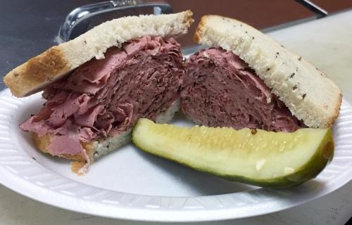 A sandwich filled with layers of pink deli meat, served with a pickle slice on a paper plate.