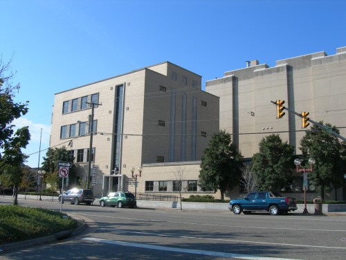 A modern building with a light-colored facade, located at a street intersection with traffic lights and trees nearby.