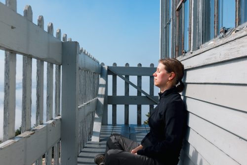 A woman sits peacefully on a balcony, eyes closed, enjoying the serene view of clouds and blue sky.