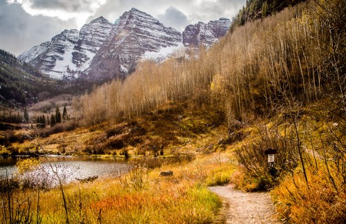 A scenic view of mountains with snow, surrounded by autumn foliage and a tranquil lake, with a winding path in the foreground.