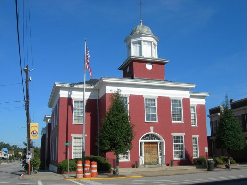Historic red brick building with a cupola, flagpole, and trees, set against a clear blue sky.