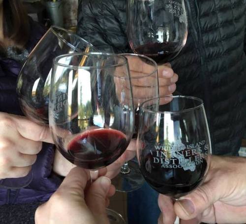 A group of hands toasting with wine glasses filled with red wine, featuring etched logos on the glasses.