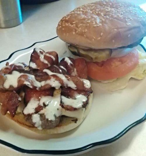A plate with a sesame seed burger topped with lettuce, tomato, and pickles, alongside a serving of bacon with sauce.