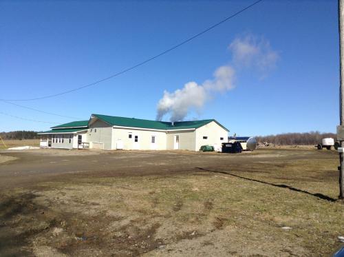 A building with a green roof emits smoke against a clear blue sky, surrounded by open land and a gravel path.