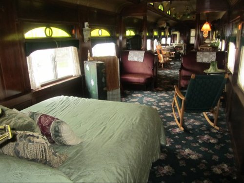 Interior of a vintage train car featuring a cozy bed, plush seating, and decorative details, with large windows.