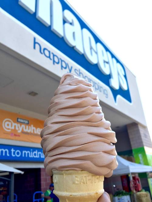 A hand holding a swirl of chocolate soft serve ice cream in a cone, with a store sign in the background.