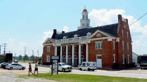 Historic brick building with a clock tower, surrounded by cars and pedestrians on a sunny day.