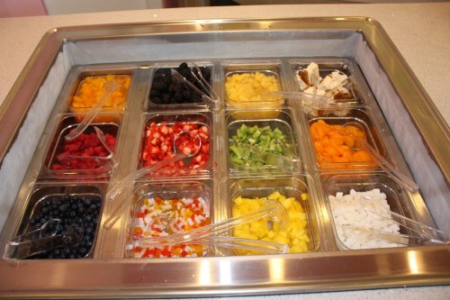 A variety of colorful fruits in clear containers, each with a spoon, arranged in a rectangular display.