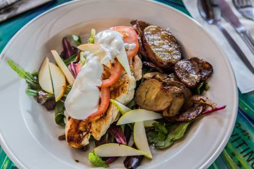 A plate of grilled chicken on a bed of mixed greens, topped with tomatoes and yogurt, alongside roasted potatoes and pear slices.