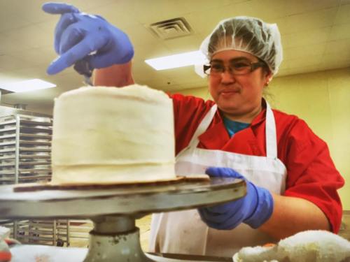A baker in a red apron and gloves decorates a white cake on a rotating stand in a kitchen.