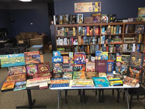 A variety of board games displayed on a table, with shelves of more games in the background.