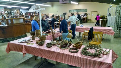 A craft fair scene with tables displaying woven baskets and people engaging in conversation in a spacious venue.