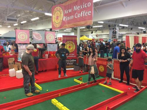 A lively event scene with people playing a game on a red carpet, surrounded by booths for New Mexico Piñon Coffee.