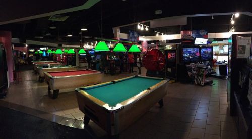 A dimly lit arcade featuring pool tables, green lamps, and various arcade games in the background.