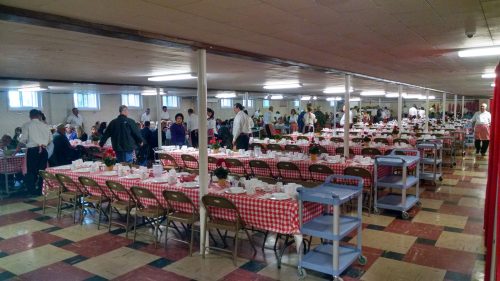 A large dining hall set with tables covered in red checkered cloth, filled with people enjoying a meal.