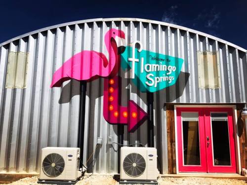 Colorful sign for "Flamingo Springs" featuring a pink flamingo and an arrow, on a metallic building with pink doors.