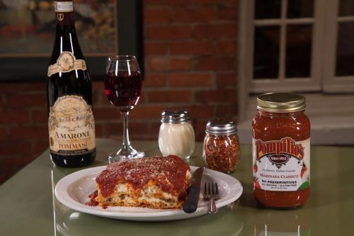 A plate of lasagna topped with marinara sauce, a bottle of wine, a glass of red wine, and condiments on a table.