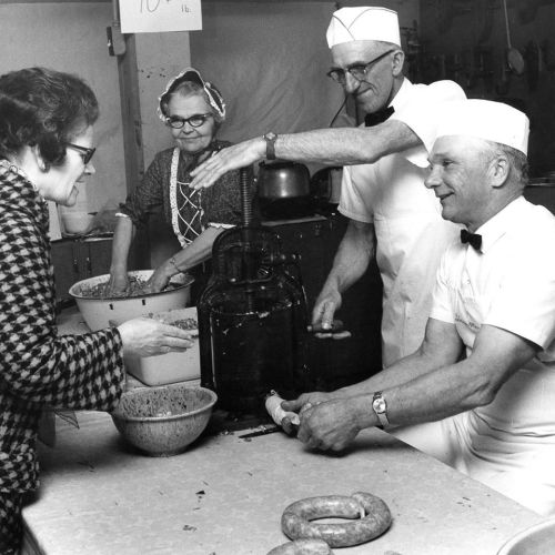 A black-and-white photo of three people making sausages in a kitchen, with one woman interacting with two men.