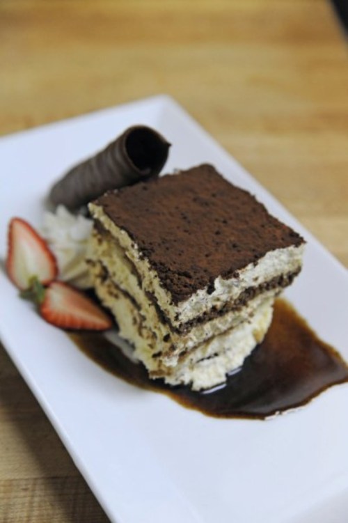A slice of tiramisu on a plate, garnished with strawberries, whipped cream, and a chocolate curl.