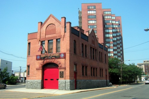 Historic brick firehouse with a red door, set against a backdrop of modern buildings and clear blue sky.