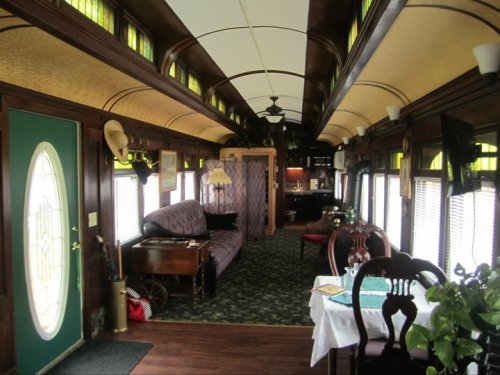 Interior of a vintage train car, featuring a cozy seating area, dining table, and kitchen, with wooden accents and green decor.