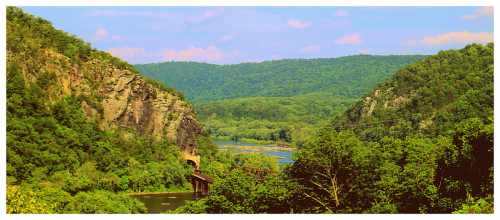 A scenic view of lush green hills and a river, with a bridge crossing through the landscape under a blue sky.