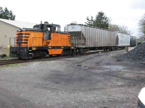An orange and black locomotive pulls a train of freight cars along a track near a building and coal pile.