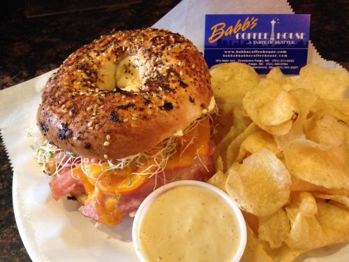 A sesame bagel sandwich with cheese, sprouts, and ham, served with a side of potato chips and a dipping sauce.