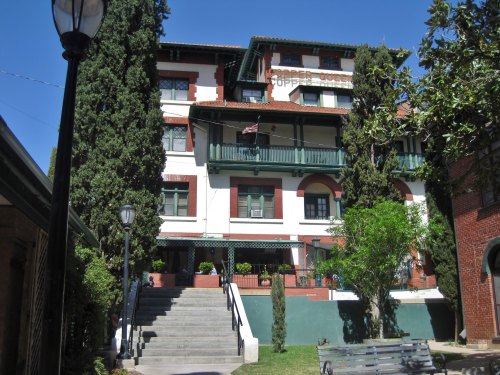Historic building with green balconies, surrounded by trees and a clear blue sky. Steps lead up to the entrance.