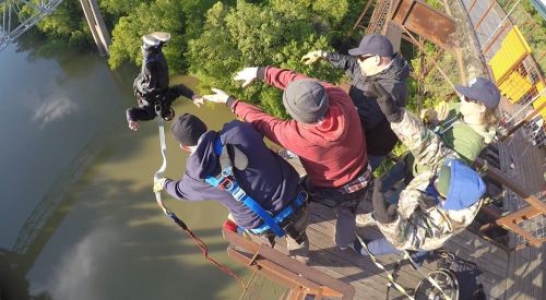 A group of people on a platform watch as someone bungee jumps off a bridge into a river below.