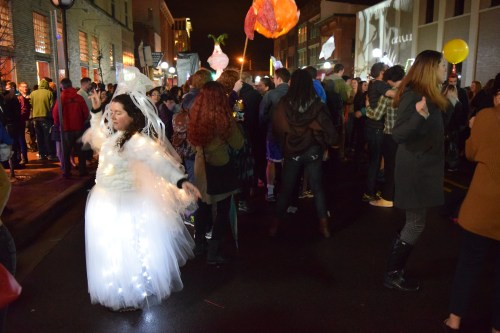 A lively street scene with a crowd celebrating, featuring a person in a glowing white dress dancing joyfully.