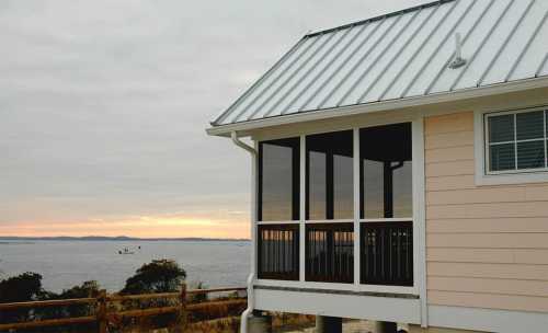 A coastal house with a metal roof and screened porch overlooks a calm sea at sunset, with soft clouds in the sky.