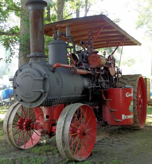 A vintage steam tractor with a large smokestack, red wheels, and a wooden canopy, set in a green outdoor environment.