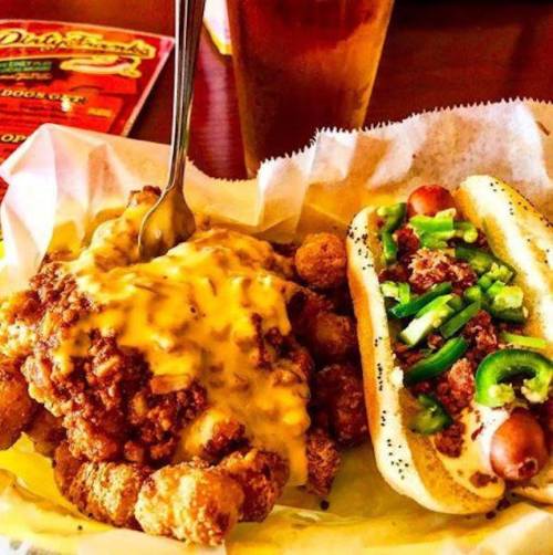 A plate of cheesy tater tots with fried chicken and a hot dog topped with bacon and jalapeños, served with a drink.