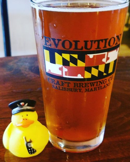 A pint of amber beer next to a yellow rubber duck wearing a police hat, on a wooden table.