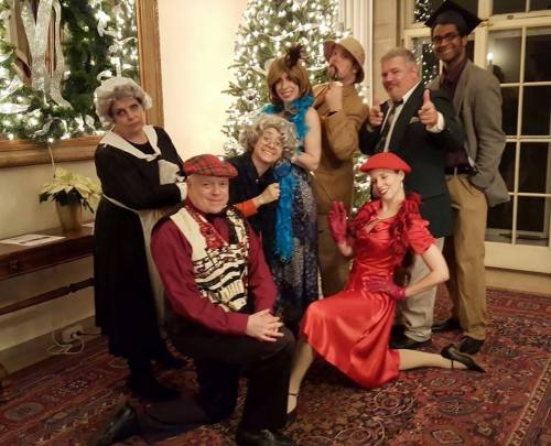 A group of eight people in festive costumes posing together in front of a decorated Christmas tree.