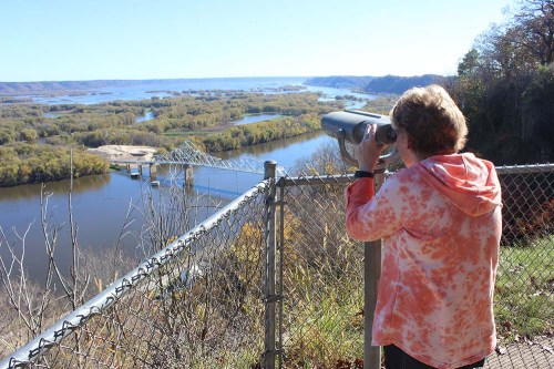 A person looks through a telescope at a scenic river view surrounded by trees and hills on a clear day.
