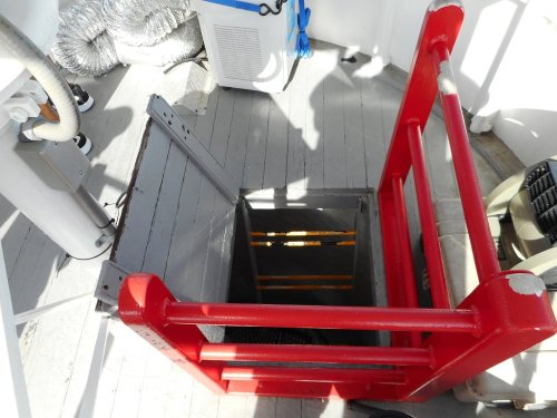 A red safety railing surrounds an open hatch on a boat, revealing a ladder leading down into the interior.