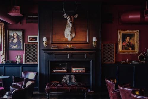 A cozy, dimly lit lounge with dark walls, vintage artwork, and a mounted deer head above a stylish fireplace.