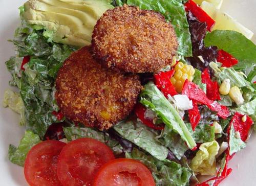 A fresh salad with greens, corn, red peppers, avocado, and two crispy patties, garnished with sliced tomatoes.