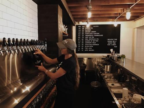 A woman fills a growler at a bar with a menu board in the background, showcasing various beers on tap.