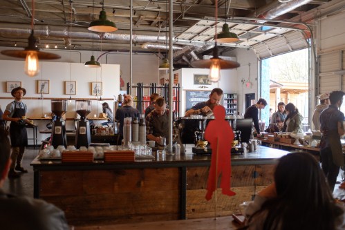 A busy coffee shop with baristas at the counter, customers seated, and industrial decor featuring hanging lights.
