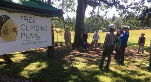 A group of people gathered under a tent with a "Tree Climbing Planet" banner in a grassy outdoor setting.